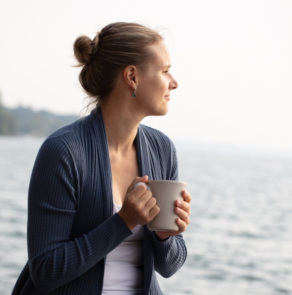 Verena founder of Yogamelia sitting by a lake holding a mug
